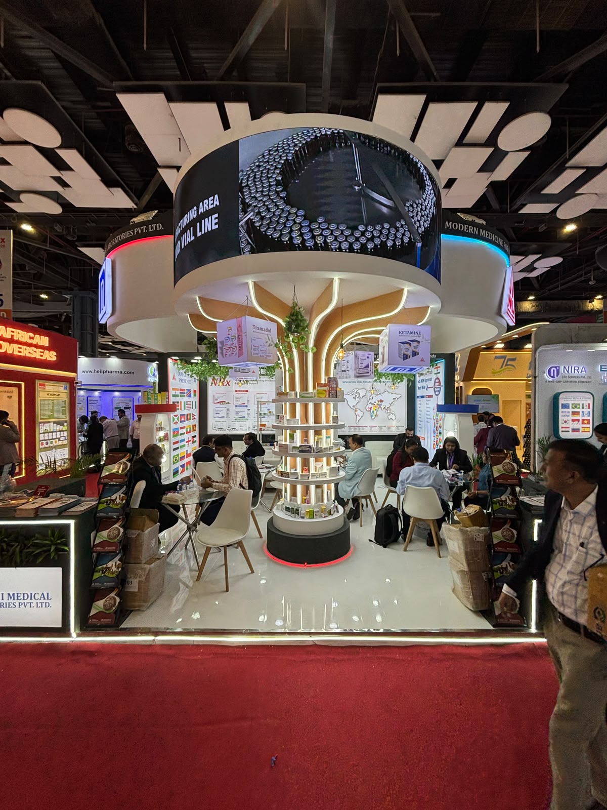 Nandani stall centrepiece — illuminated tree-shaped column with circular product shelves and a curved LED screen showing the vial line