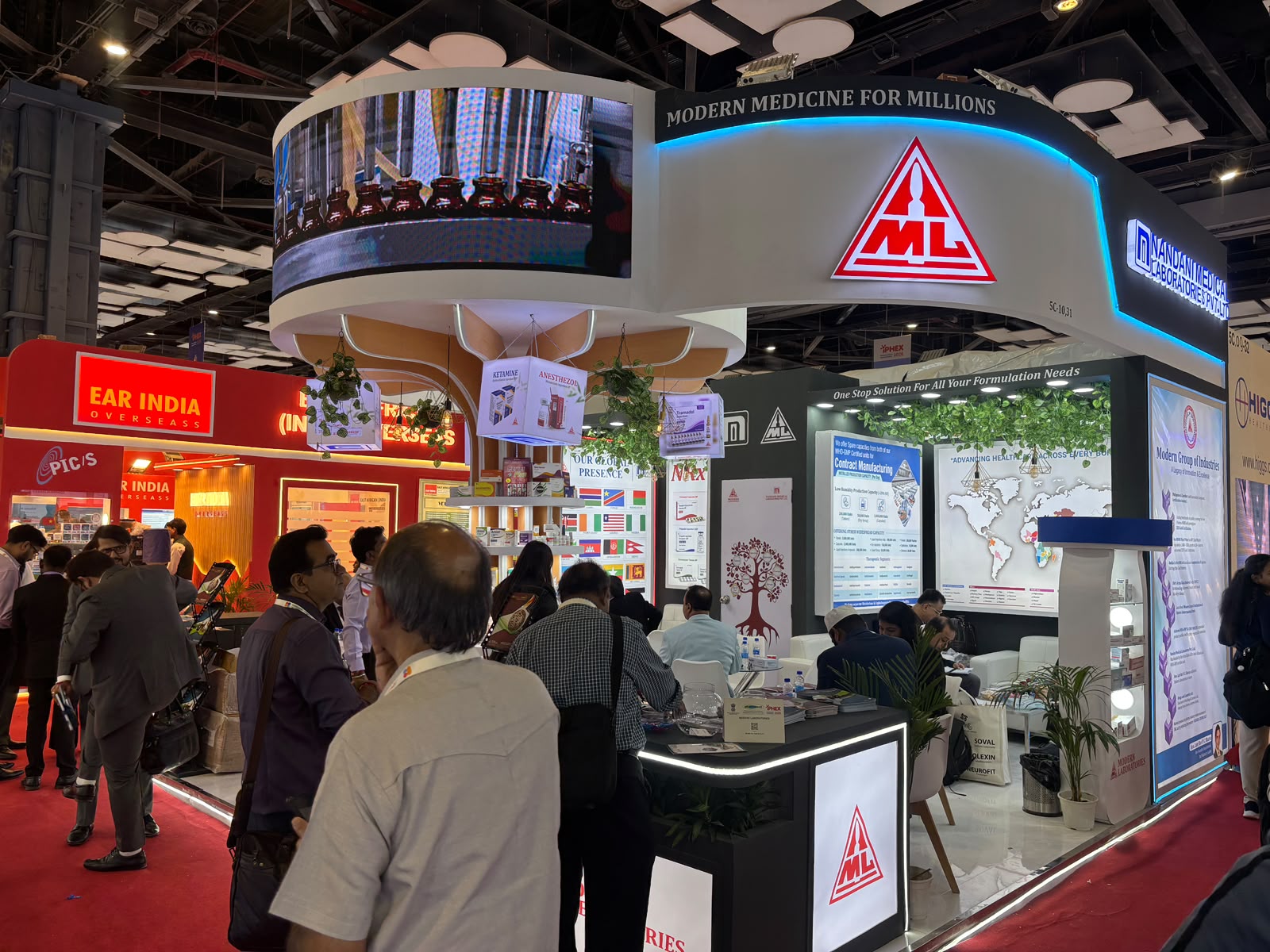 Nandani stall front view — fascia LED screen playing the amber-vial production line, visitors at the reception desk